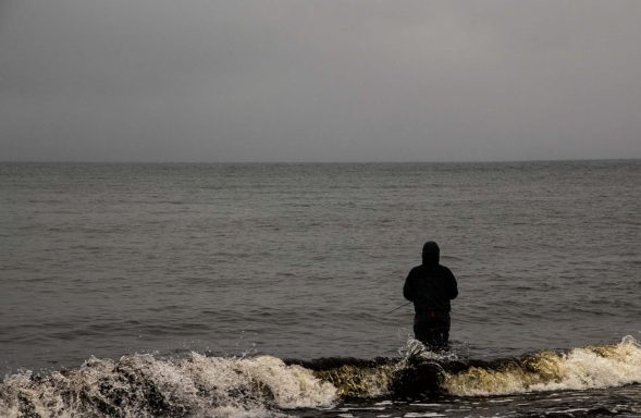 En person står i vattnet vid en grå strand under molnigt väder.