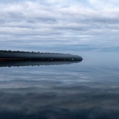 Lugnt vatten med spegelbild av en dimmig himmel och skog på horisonten.