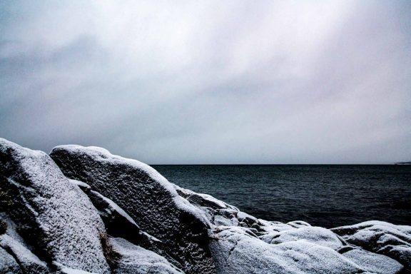 Klippor täckta av snö vid en mörk havshorisont under en molnig himmel.