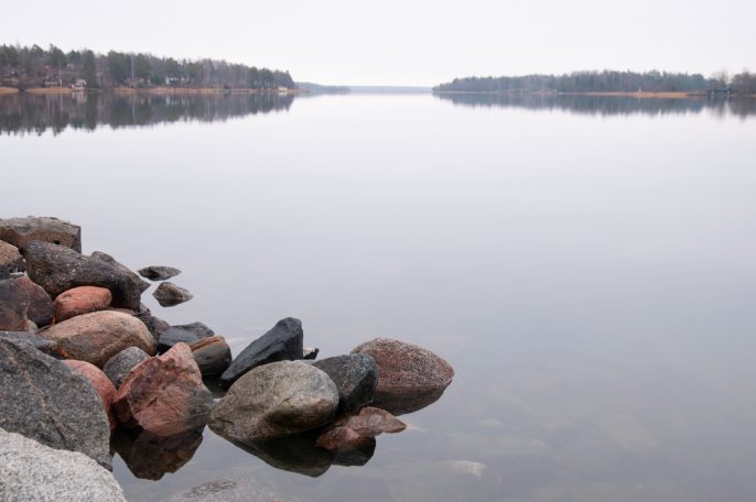 Stenar vid en spegelblanka sjö med skogslandskap i bakgrunden.