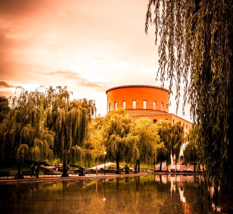 Rund byggnad vid vattnet omgiven av träd och en orangeaktig himmel.