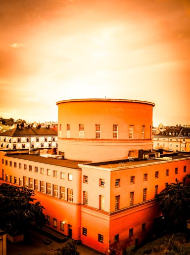 Rund orange byggnad med ett orange tak, omgiven av gröna träd och en ljus himmel.