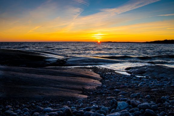 Solnedgång över havet med reflektioner på vattnet och klippor i förgrunden.